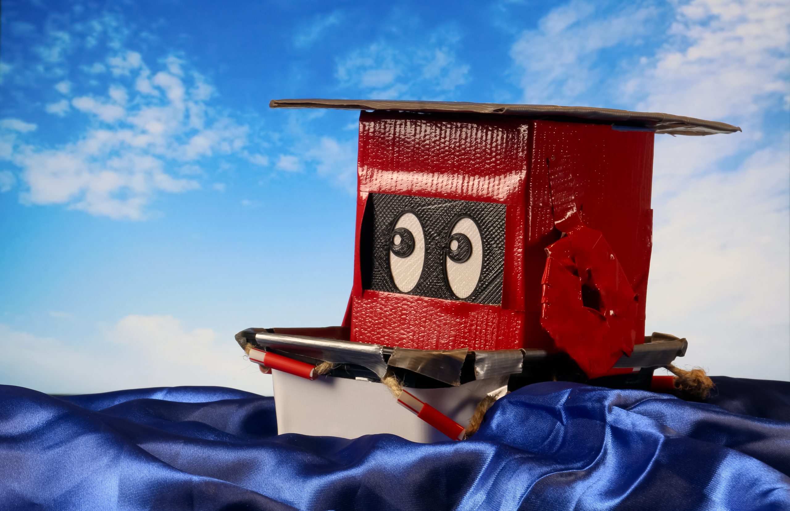 Recyclobot Toy Tim Brody (TB-12) The Tugboat sitting on blue wavy fabric and a blue and white cloud sky background